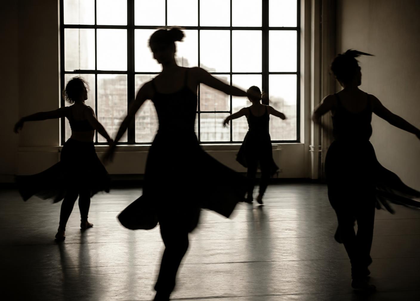 Dancers rehearsing in a bare studio, silhouetted against a large window